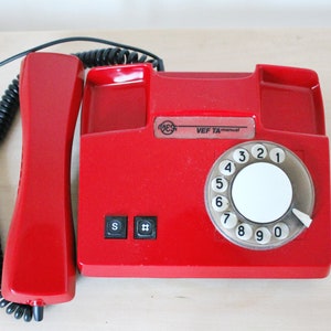 Puede incluir: Un teléfono rojo de disco con un cable negro en espiral y un disco blanco. El teléfono tiene una etiqueta que dice "VEF TA-manual".