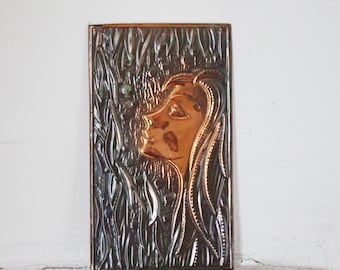 May include: A copper wall plaque with a raised relief design of a woman's face with long hair. The background is a textured design of leaves.