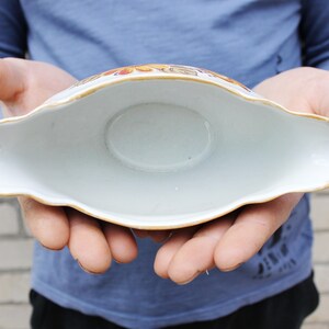 May include: A white ceramic gravy boat with a gold rim and floral pattern. The boat is empty and being held by a person's hands.