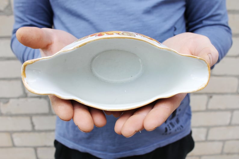 May include: A white ceramic gravy boat with a gold rim and floral pattern. The boat is empty and being held by a person's hands.