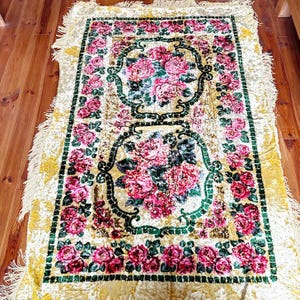 May include: A rectangular rug with a cream background and a large floral pattern. The central design features pink and red flowers with green foliage, framed by a green border. The rug has a fringed edge and is likely made of a soft material.