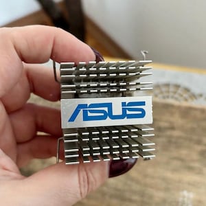 Peut inclure: Un composant informatique ASUS argenté avec un logo bleu. Le composant comporte une série d'ailettes parallèles pour la dissipation de la chaleur. Le logo ASUS est imprimé sur une plaque argentée.