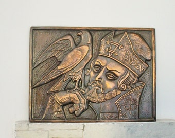 May include: A bronze wall plaque depicting a man with a beard and a crown, holding a falcon. The plaque has a textured surface and intricate details.