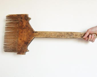 Könnte beinhalten: Ein Holzkamm mit langem Griff, der zum Kämmen von Wolle verwendet wird. Der Kamm hat einen breiten, flachen Kopf mit einer Reihe von Zähnen auf einer Seite. Der Griff ist glatt und abgerundet.