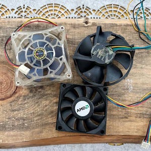 Peut inclure: Trois ventilateurs de refroidissement d'ordinateur sont présentés sur une surface en bois. Un ventilateur est transparent avec des pales bleues, un autre est noir et le troisième est noir avec un logo AMD. Chaque ventilateur a des fils colorés attachés.