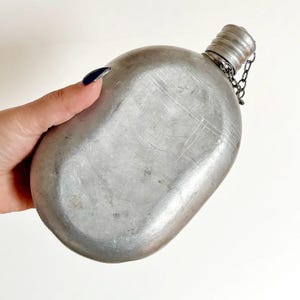 May include: A vintage, oval-shaped canteen made of silver-colored metal. The canteen has a screw-on cap attached by a chain. The metal shows signs of wear, with scratches and a slightly aged appearance. The canteen is held by a hand.