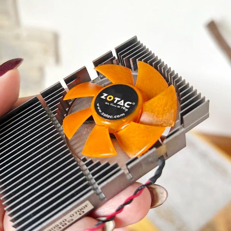 Pode incluir: Um cooler de placa gr&aacute;fica de computador com uma ventoinha de l&acirc;minas laranja e um log&oacute;tipo ZOTAC preto. O cooler tem um dissipador de calor de metal prateado com aletas paralelas. A ventoinha foi concebida para dissipar o calor.