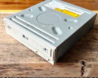 May include: A gray LG DVD-ROM drive with a silver top and a front panel with the LG logo. The drive has a disc tray and a small button labeled "Secure Disc Open". The drive is on a wooden surface.