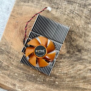 Pode incluir: Uma ventoinha de computador com p&aacute;s laranja e um centro preto com o log&oacute;tipo ZOTAC. A ventoinha est&aacute; montada num dissipador de calor prateado com aletas pretas. Um fio vermelho e preto sai da ventoinha.