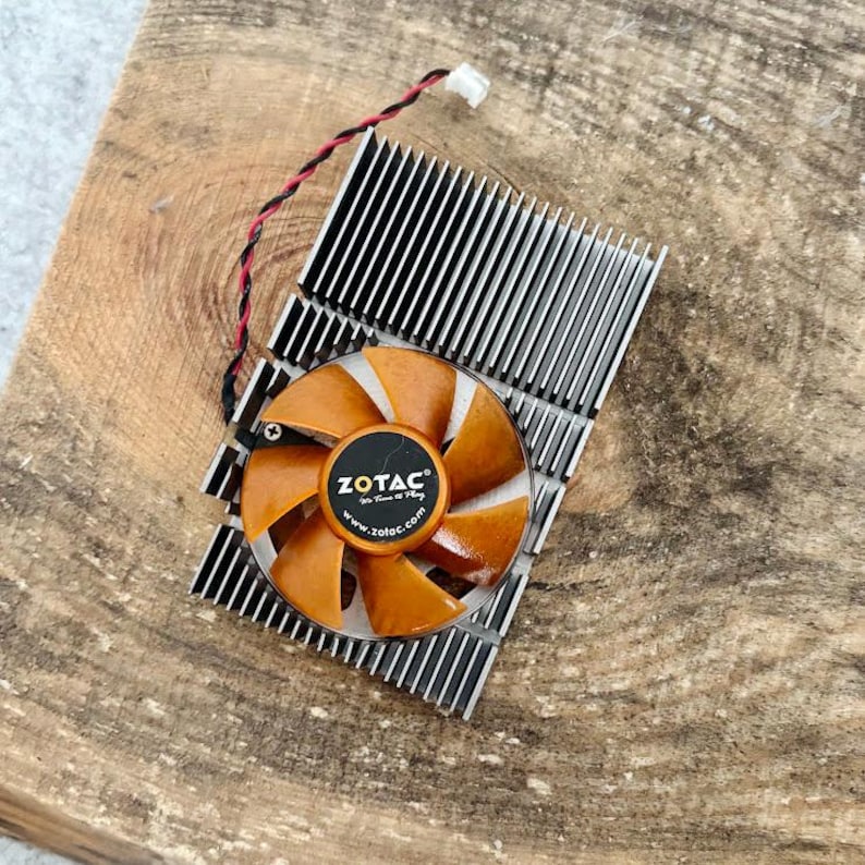 Pode incluir: Uma ventoinha de computador com p&aacute;s laranja e um centro preto com o log&oacute;tipo ZOTAC. A ventoinha est&aacute; montada num dissipador de calor prateado com aletas pretas. Um fio vermelho e preto sai da ventoinha.