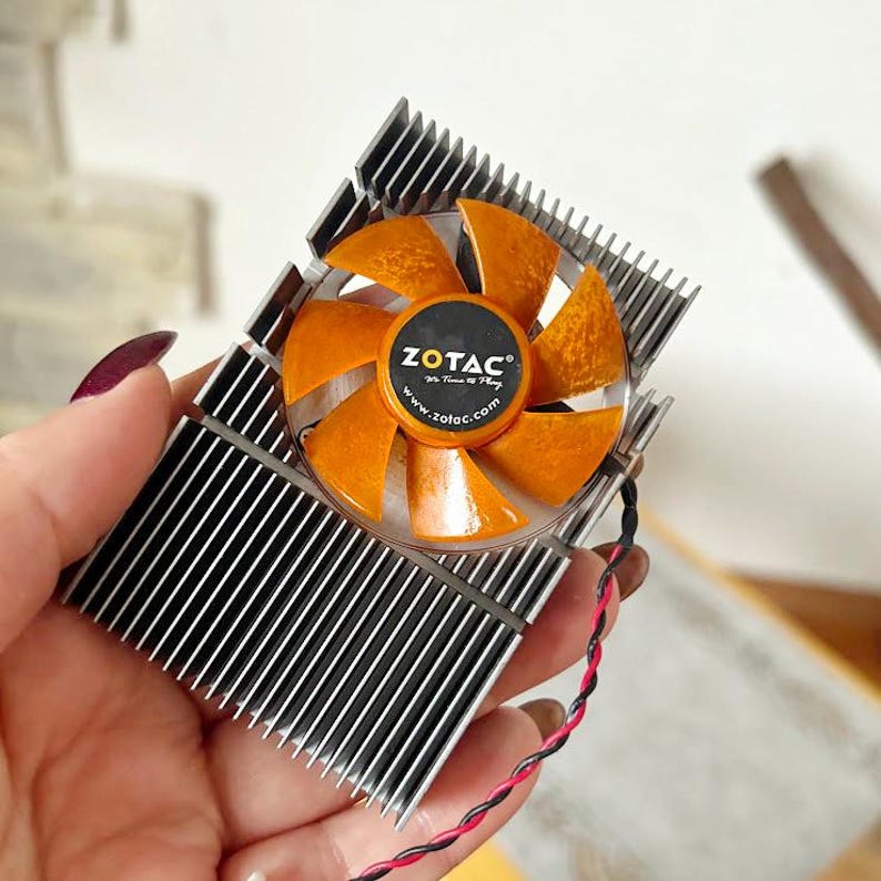 Pode incluir: Um cooler de placa gr&aacute;fica de computador com uma ventoinha com l&acirc;minas laranja e um centro preto com o logotipo e site da ZOTAC. O cooler tem um dissipador de calor prateado com aletas verticais. Um fio preto e vermelho se estende do cooler.