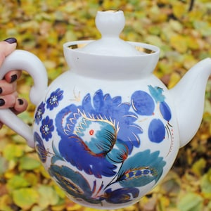 Puede incluir: Tetera de porcelana blanca con un diseño floral azul y dorado. La tetera tiene una tapa y un pico. El fondo es una imagen borrosa de hojas de otoño.