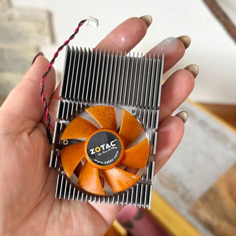 Pode incluir: Um cooler de placa gr&aacute;fica de computador com um dissipador de calor prateado e uma ventoinha laranja. A ventoinha tem a marca "ZOTAC" impressa. O cooler tem um fio preto e vermelho.