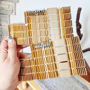 Puede incluir: Dos disipadores de calor de metal dorado para computadora con estructuras de aletas verticales. Cada disipador cuenta con una placa rectangular negra con el texto blanco GIGABYTE impreso. Los componentes tienen un acabado metálico y diseño estriado.