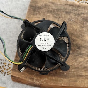 May include: A black CPU cooler with a fan and a circular label that reads "CPU COOLER Ok TET DC 12V Made in China CE". The fan has multiple blades and is connected to wires with yellow, green, and blue insulation.
