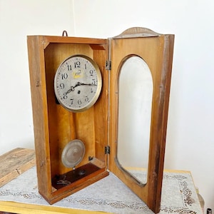 May include: A vintage wooden wall clock with an open door. The clock face is silver with black numbers and hands. A pendulum and other clock components are visible inside the wooden case. The clock's door has a glass panel.