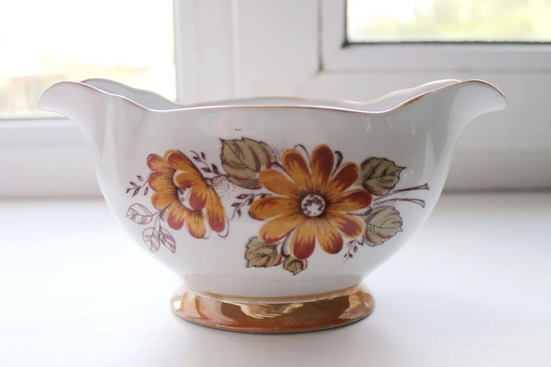 May include: A white ceramic gravy boat with a gold rim and floral design featuring brown and orange flowers.