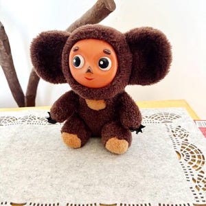 May include: A plush brown toy with large ears and a round, orange face. The toy has a brown hood, a tan chest patch, and black claws. It is sitting on a gray surface.