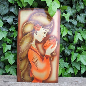 May include: A vintage wall art piece featuring a woman with long brown hair wearing a red dress and holding a small orange animal. The background is a gold metallic color.