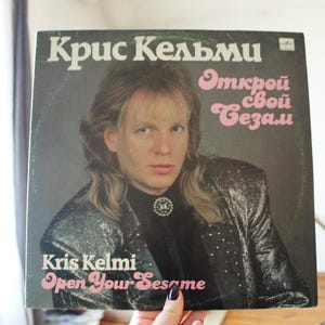 May include: A vinyl record cover featuring Kris Kelmi, a man with long blonde hair, wearing a black jacket with silver sequins. The cover is black with pink text that reads "Открой свой Сезам" and "Open Your Sesame" in English.