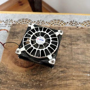 May include: A black and silver computer fan with a circular grill design. The fan has a central label with the brand name "TITAN" and technical specifications. The fan is secured with four screws and has wires extending from it.