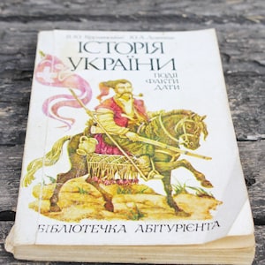 Op de afbeelding: Een vintage boekomslag met een vergeelde achtergrond en een kleurrijke illustratie van een man te paard. De titel van het boek is "Історія України", wat vertaalt naar "Geschiedenis van Oekraïne". Het boek maakt deel uit van een serie genaamd "Бібліотечка абітурієнта", wat vertaalt naar "Bibliotheek voor aanvragers".