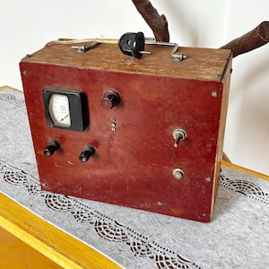 May include: A vintage rectangular electronic device housed in a wooden box with a dark red front panel. It features a black analogue gauge, a brown rotary knob, two black terminals, a metal toggle switch, a small button, and a black power plug on top.
