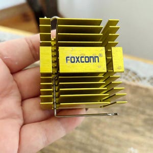 May include: A yellow metal heat sink with the text "FOXCONN" in blue. The heat sink has multiple parallel fins and a metal wire clip. The object is held in a hand.