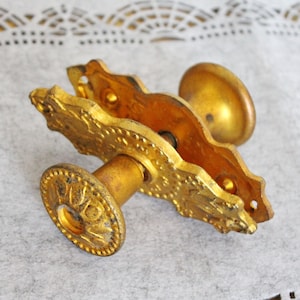 May include: A gold-toned metal door handle with an ornate design. The handle has a round knob on one end and a flat plate with a keyhole on the other end.