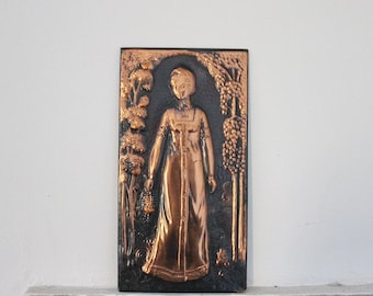 May include: A copper wall hanging featuring a woman in a long dress standing in a garden setting. The woman is surrounded by trees and vines. The wall hanging is rectangular and has a dark brown background.