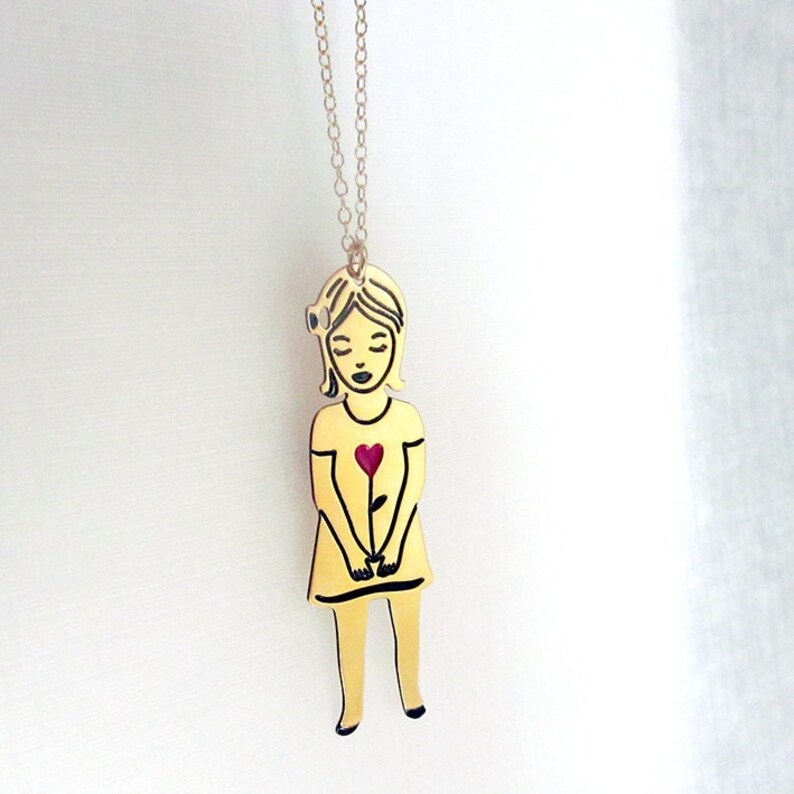 Op de afbeelding: Gouden ketting met een hanger van een meisje dat een rode hartvormige bloem vasthoudt. Het meisje draagt een jurk en heeft een bloem in haar haar.