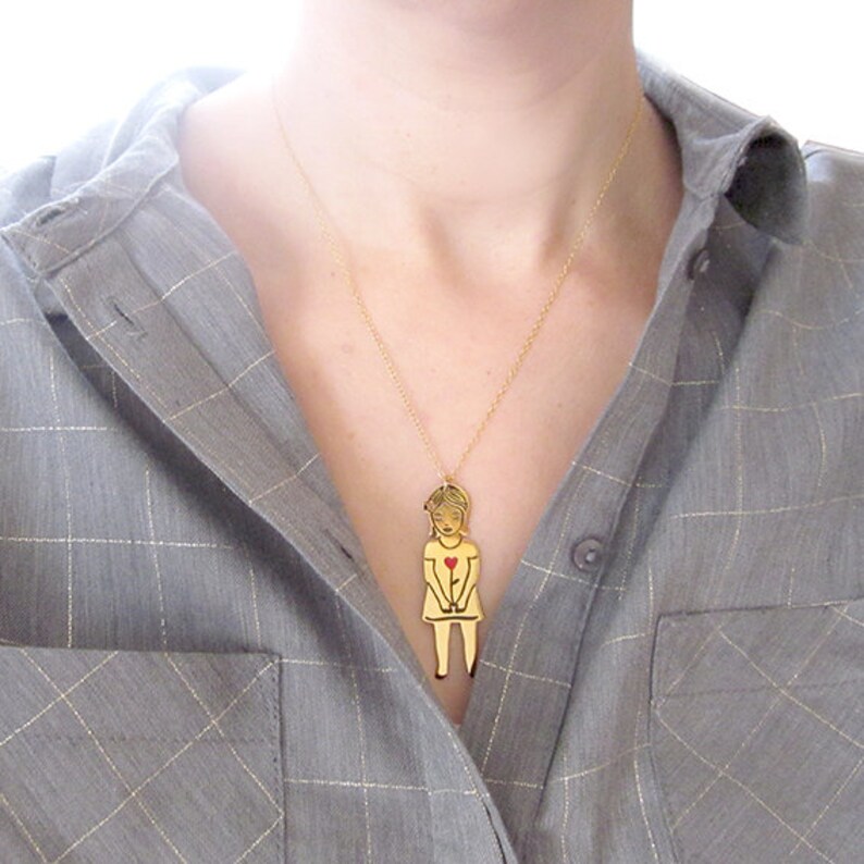 Op de afbeelding: Een gouden ketting met een hanger met een gestileerde illustratie van een persoon die een rood hart vasthoudt. De hanger is gemaakt van metaal en heeft een glanzende afwerking.