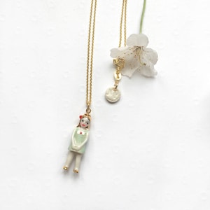 Op de afbeelding: Twee gouden kettingen met porseleinen hangers. Een hanger is een klein meisjesfiguurtje dat een groene jurk en rode schoenen draagt. De andere hanger is een witte cirkel.