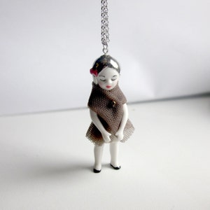 Op de afbeelding: Een porseleinen poppenhanger met een bruine tule jurk en een zilveren ketting. De pop heeft een wit gezicht met zwarte ogen en roze lippen.