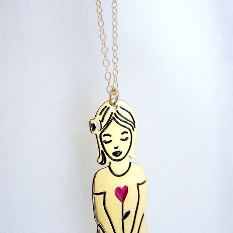 Op de afbeelding: Gouden ketting met een hanger van een vrouw met een rood hart op haar borst. De vrouw is zwart omlijnd en heeft haar ogen gesloten.