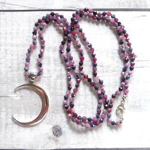 May include: A beaded necklace with a crescent moon pendant. The necklace features small, faceted beads in shades of pink, purple, and dark blue. The pendant is a polished silver color. The necklace is displayed on a light wood surface.
