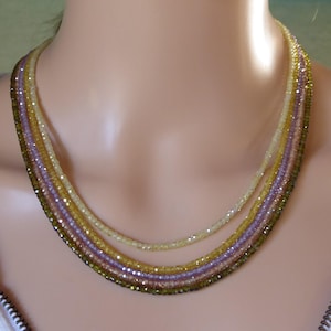 May include: A multi-strand necklace featuring four distinct layers of small, faceted, translucent beads. The strands are arranged in a cascading design, showcasing a color palette of pale yellow, golden yellow, soft lavender, and deep olive green.