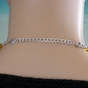 May include: Close-up of a silver-toned necklace clasp and extender chain. The chain is attached to a necklace with yellow and green beads. The necklace is around a person's neck, with a black top visible.