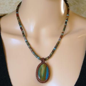 May include: A handcrafted necklace featuring a pendant with a blue, green, and brown stone. The pendant is encased in a brown woven setting and hangs from a beaded strand with various earth-tone colors. The necklace is displayed on a mannequin.