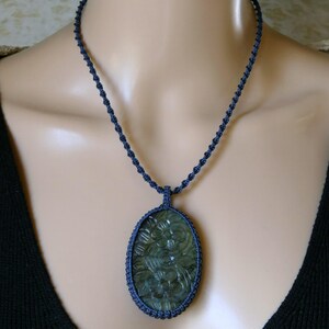 May include: A dark blue macrame necklace with an oval pendant. The pendant features a carved floral design in shades of green and is encased in a macrame border. The necklace is displayed on a mannequin.