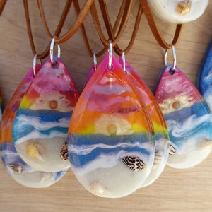 May include: A set of colorful resin pendants with a beach sunset theme. Each pendant features a layered design with blue, white, and yellow colors, representing the ocean, sand, and sunset. The pendants are attached to brown leather cords.