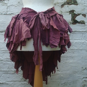 Peut inclure: Vêtement en tissu bordeaux superposé, possiblement un châle ou une jupe, présenté sur un mannequin. Le tissu semble vieilli ou effiloché intentionnellement, avec une fermeture à nouer à l'avant. Fond de mur en briques blanches patinées.
