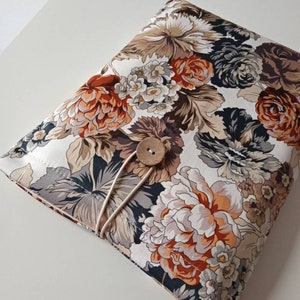 May include: A floral-patterned book cover featuring large, stylized flowers in shades of orange, brown, cream, and gray. The cover is secured with a wooden button and an elastic loop. The overall design is reminiscent of vintage fabric.