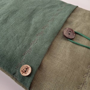 May include: Close-up of a two-toned linen pouch. The top flap is a deep green, stitched with dark thread, and secured with a wooden button. The bottom section is a lighter olive green. A second button and green elastic cord are visible.