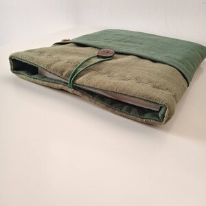 May include: A green and tan laptop sleeve made of textured fabric. The sleeve has a button closure and a green elastic band. The top flap is a darker shade of green. The sleeve is designed to protect a laptop.