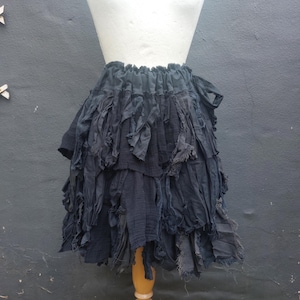 Puede incluir: Falda negra de varias capas hecha de tela rasgada, exhibida en un maniquí blanco. La falda tiene una cintura fruncida y una apariencia desgastada y texturizada. La falda mide aproximadamente 46 cm de largo.