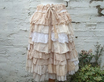 May include: A tiered skirt in shades of beige and white. The skirt has a ruffled design with layers of fabric. A beige drawstring is visible at the waist. The skirt is set against a textured white brick wall.