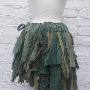 Peut inclure: Une jupe composée de multiples couches de bandes de tissu vert olive et vert forêt aux bords effilochés et bruts. Le vêtement présente une taille élastique avec un lien de serrage latéral, créant un aspect texturé et superposé sur un mannequin.