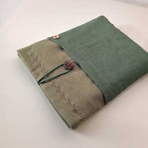 May include: A folded, olive green and dark green linen case with a button closure. The case has a decorative stitched pattern and a green elastic loop with a wooden button. The case is on a white surface.