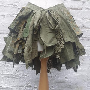 Peut inclure: Une jupe superposée vert olive avec une taille à nouer, composée de divers morceaux de tissu incluant du coton uni, des textiles à motifs et des panneaux de dentelle au crochet complexes, créant un aspect texturé et multidimensionnel.